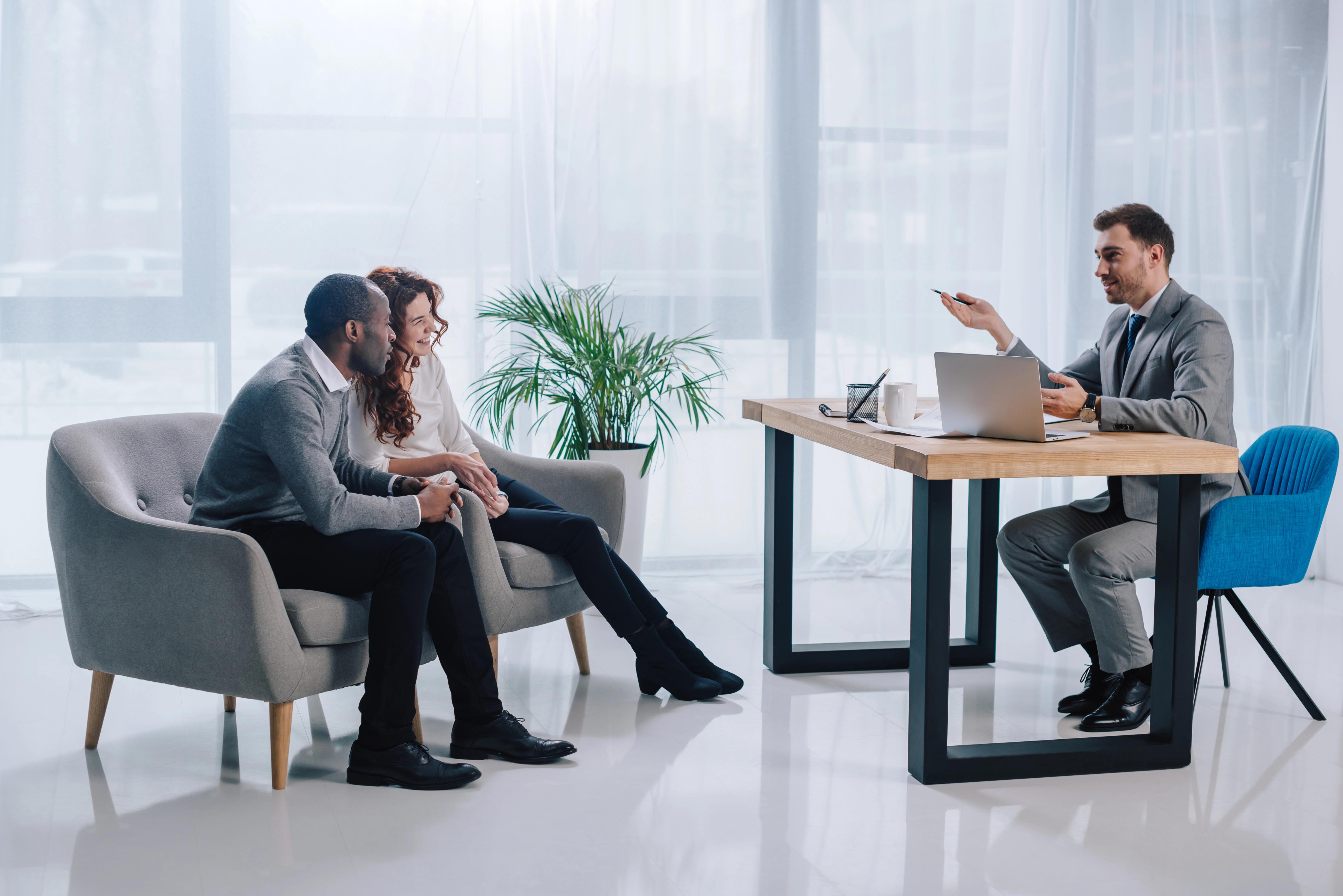 Interracial couple sitting in armchairs and estate agent at table with laptop Interracial couple sitting in armchairs and estate agent at table with laptop
